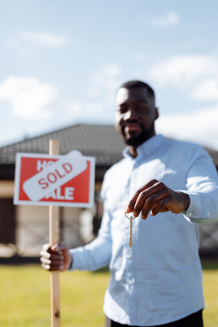 Smiling black real estate agent holding key and sold sign on sunny day outdoors.